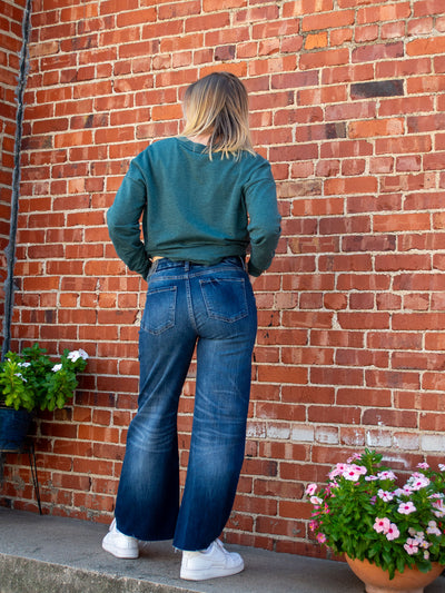 A model wearing a dark wash, faded, wide leg jean with a raw hem. The model has it paired with a teal v-neck top.