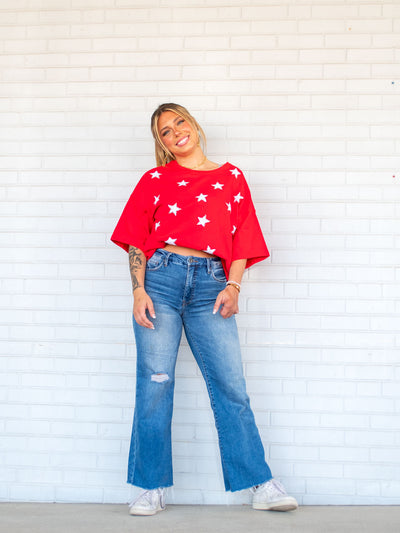 A model wearing a pair of wide leg, flared jeans with a hole on the knee. The model has it paired with a red tee with stars and white sneakers.