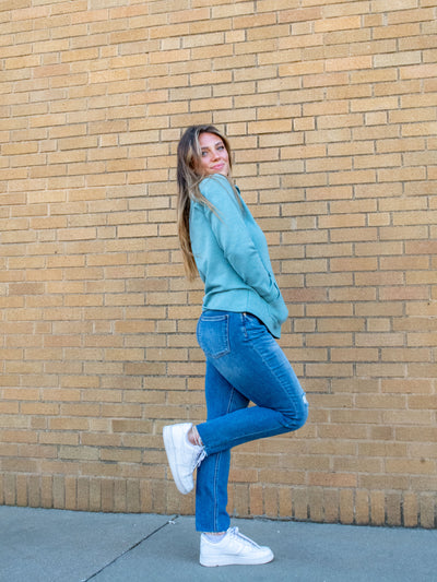 A model wearing a pair of medium wash jeans with distressing at the knees. The model has them paired with a teal pullover and white sneakers.