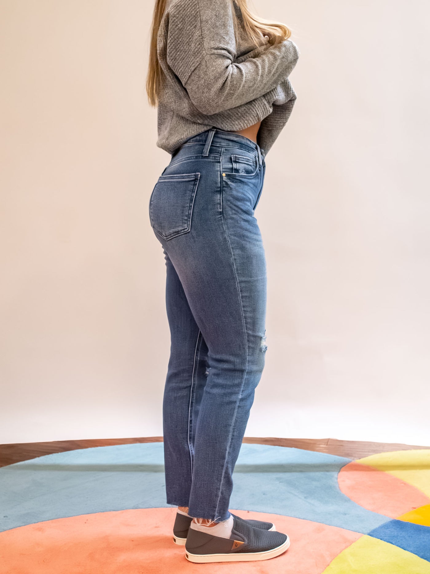 A model wearing a pair of medium washed jeans with holes and distressing and an unfinished hem. The model has it paired with a gray sweater and gray loafers.