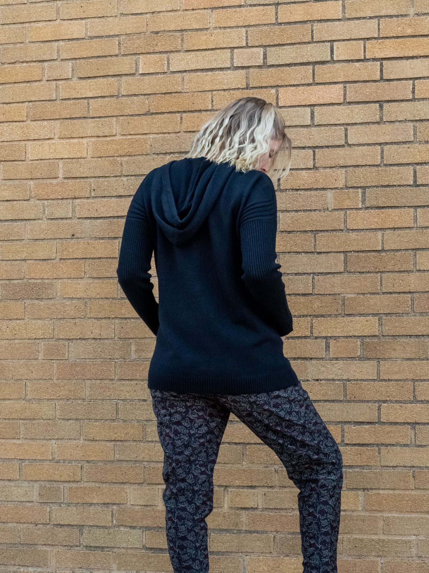 A model wearing a black hooded sweater with drawstrings and pockets. The model has it paired with a black, gray, and red printed jogger.