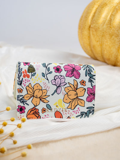 A white card and cash holder with a yellow, pink, teal, yellow, and purple flower design on it. The wallet folds and has a gold snap and 3 pockets inside. 