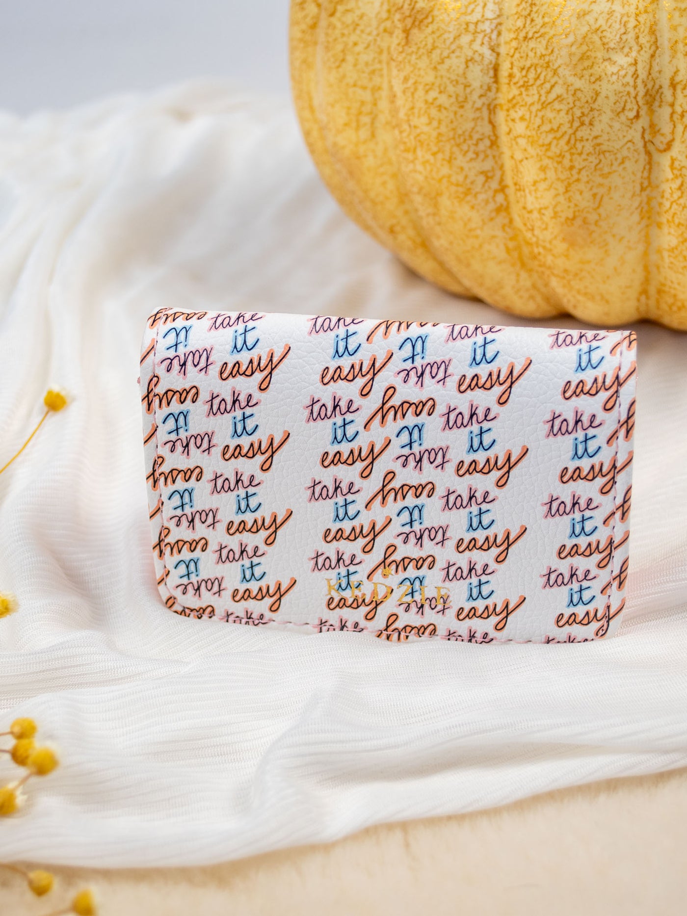 A white card and cash holder with a white base and "take it easy" written pattern on it. The wallet folds and has a gold snap and 3 pockets inside. 
