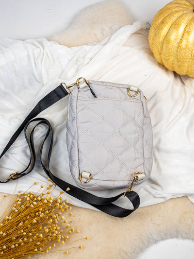A gray colored quilted bag with three gold zipper pockets and black adjustable straps.
