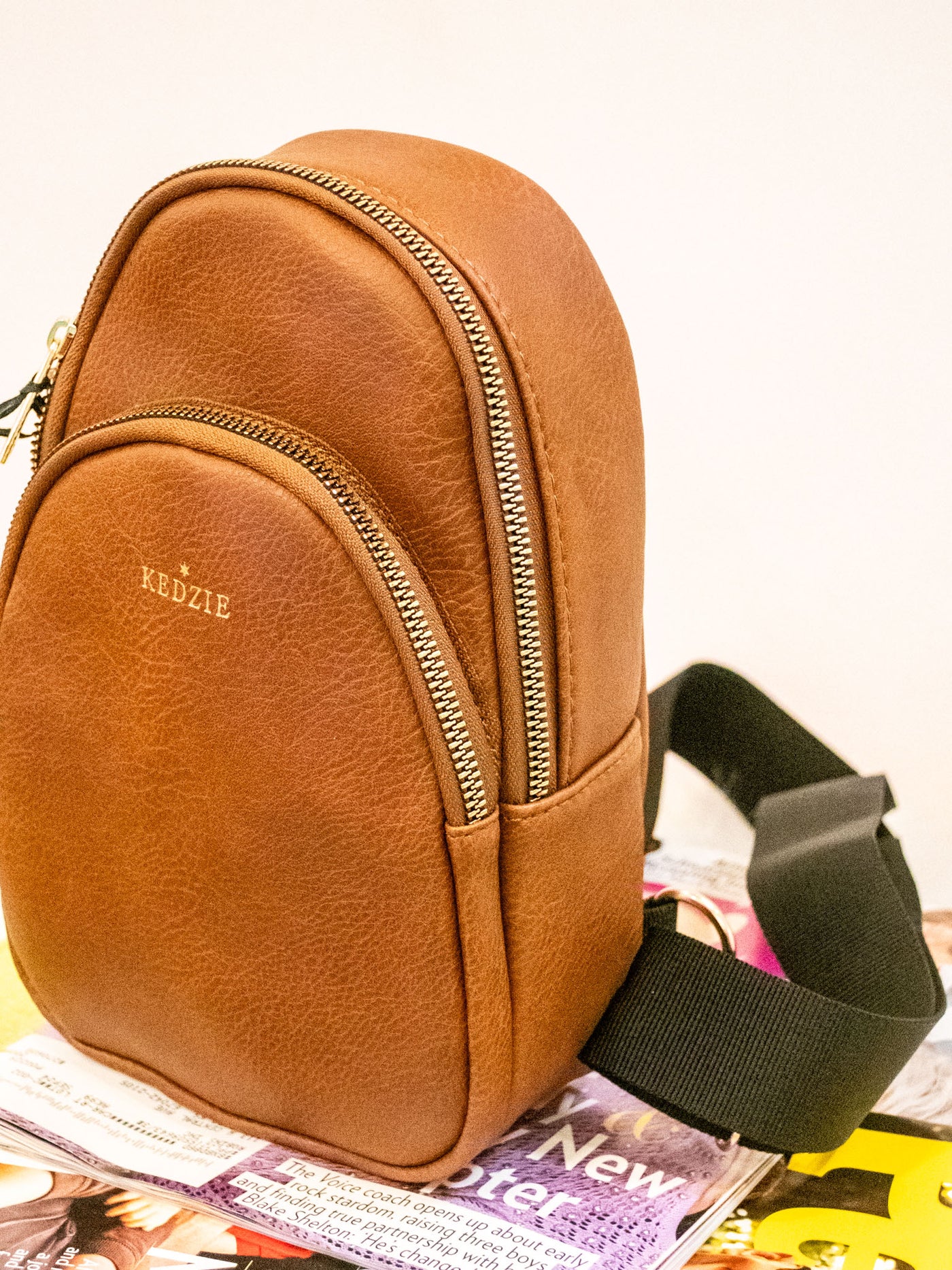 A brown vegan leather sling bag with two zipper pockets and a black strap.