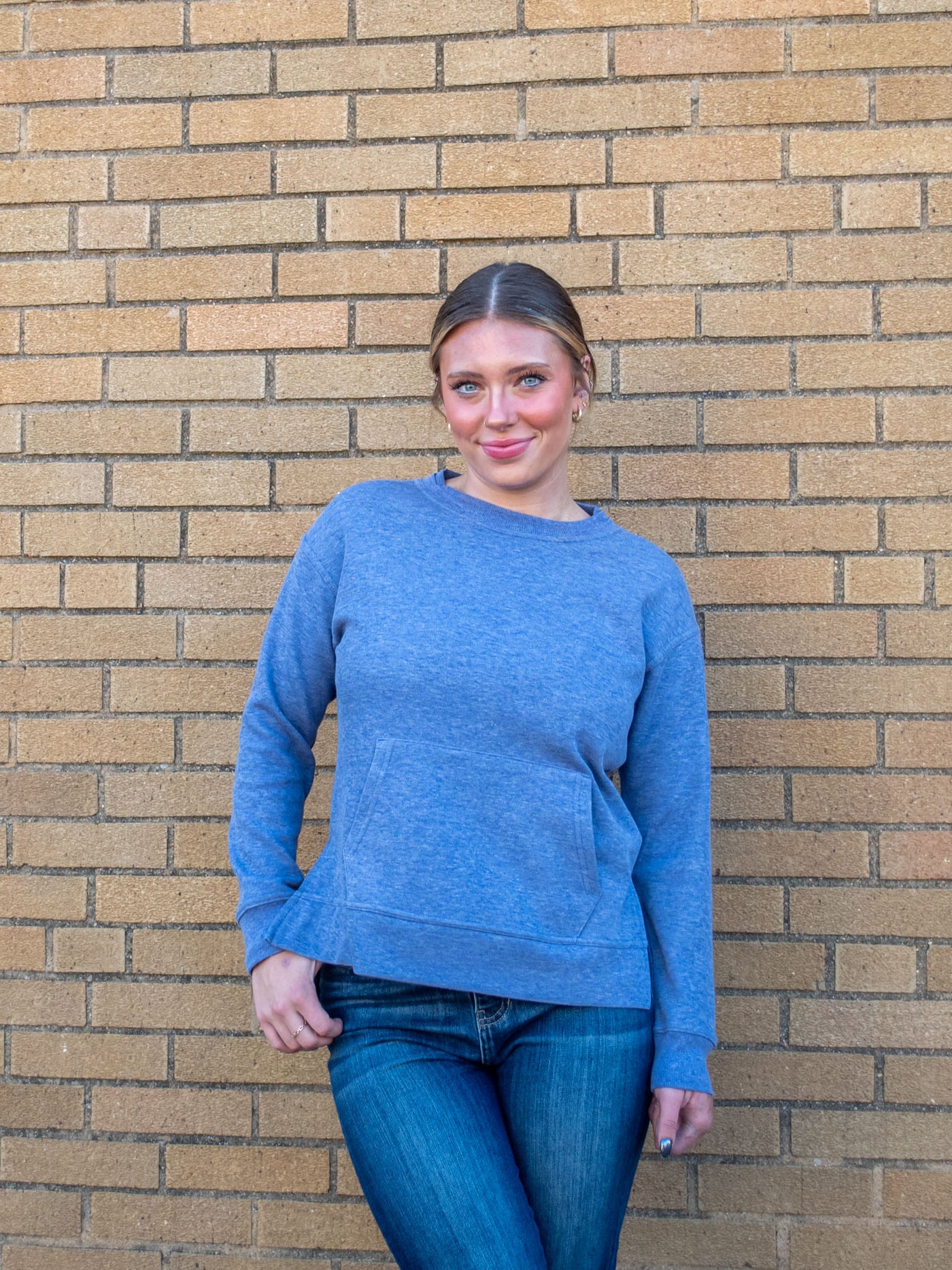 A model wearing a blue crewneck sweatshirt with a front pocket and side slits. The model has it paired with a dark wash jean.