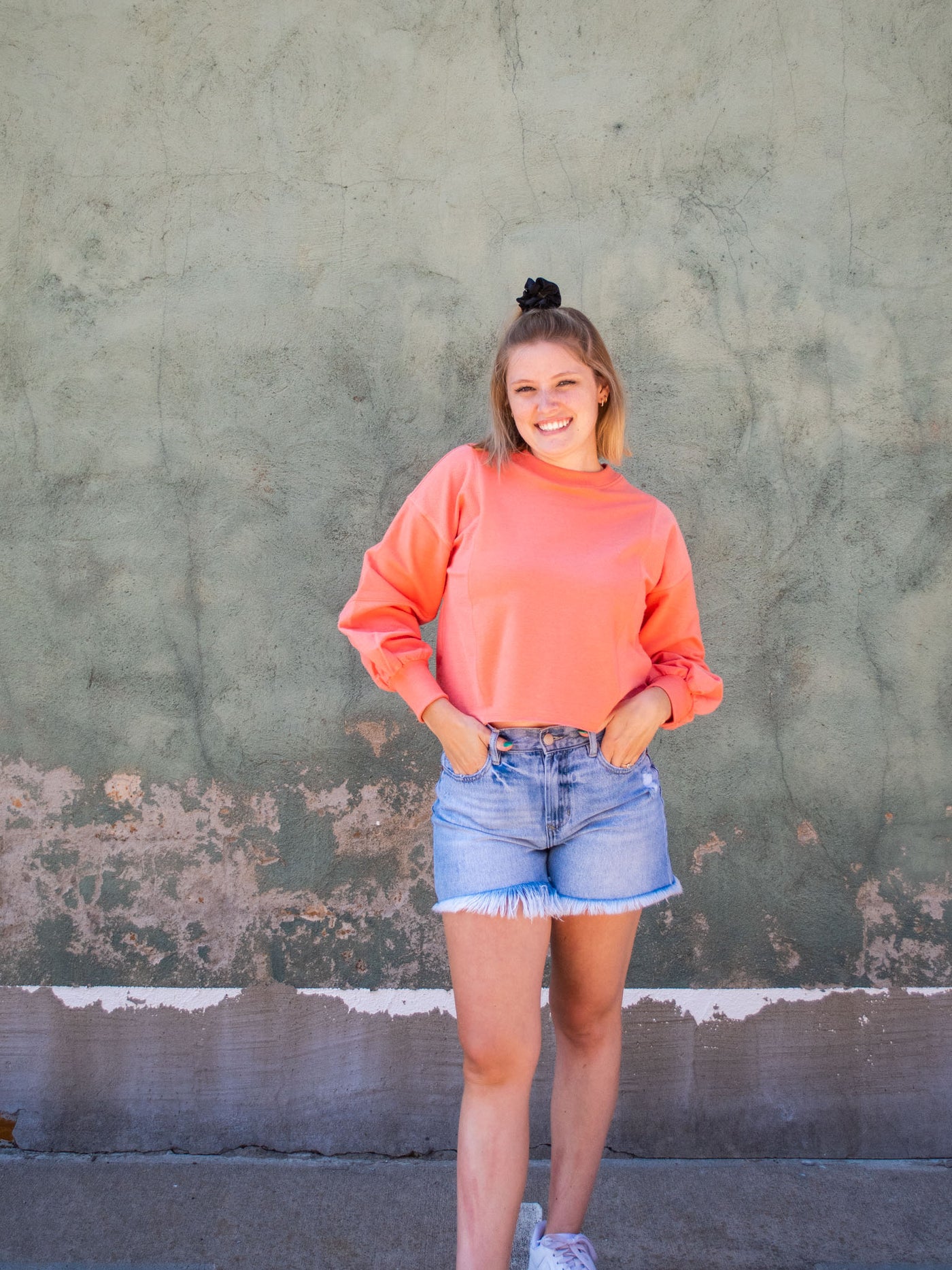 A model wearing a coral colored cropped sweatshirt with arm stitching details. She has it on with light washed, frayed jean shorts