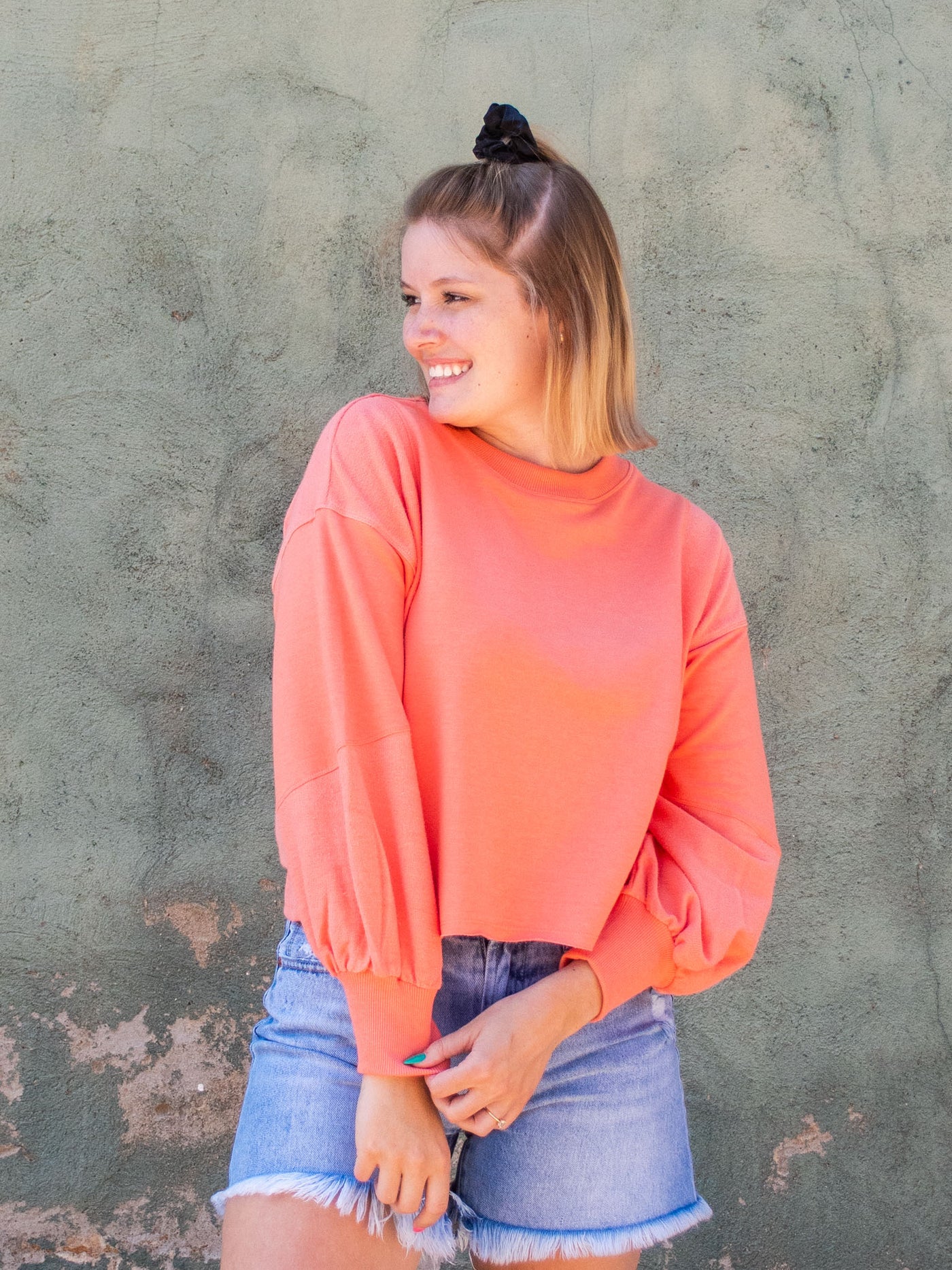 A model wearing a coral colored cropped sweatshirt with arm stitching details. She has it on with light washed, frayed jean shorts