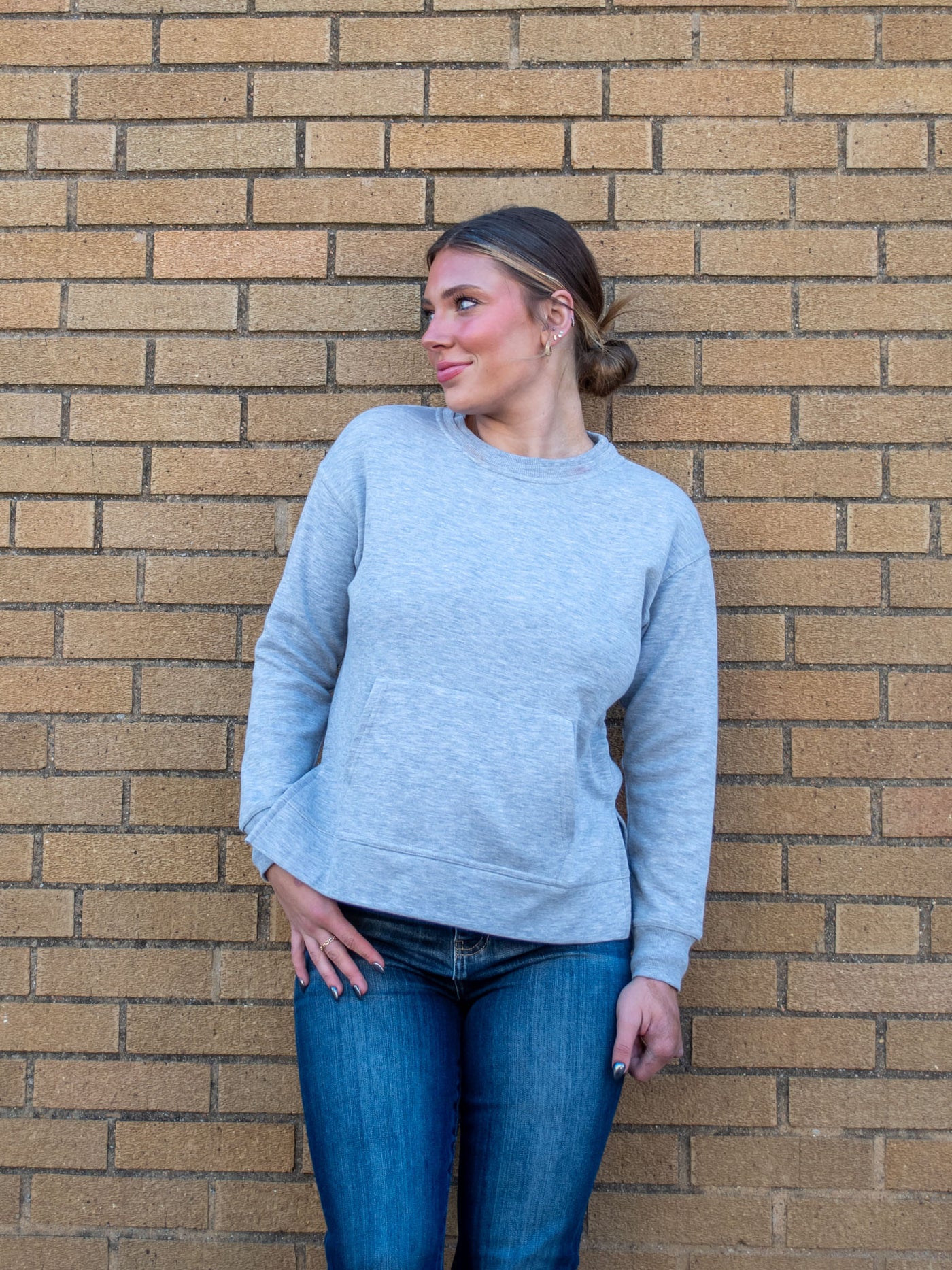 A model wearing a light gray crewneck sweatshirt with a front pocket and side slits. The model has it paired with a dark wash jean.