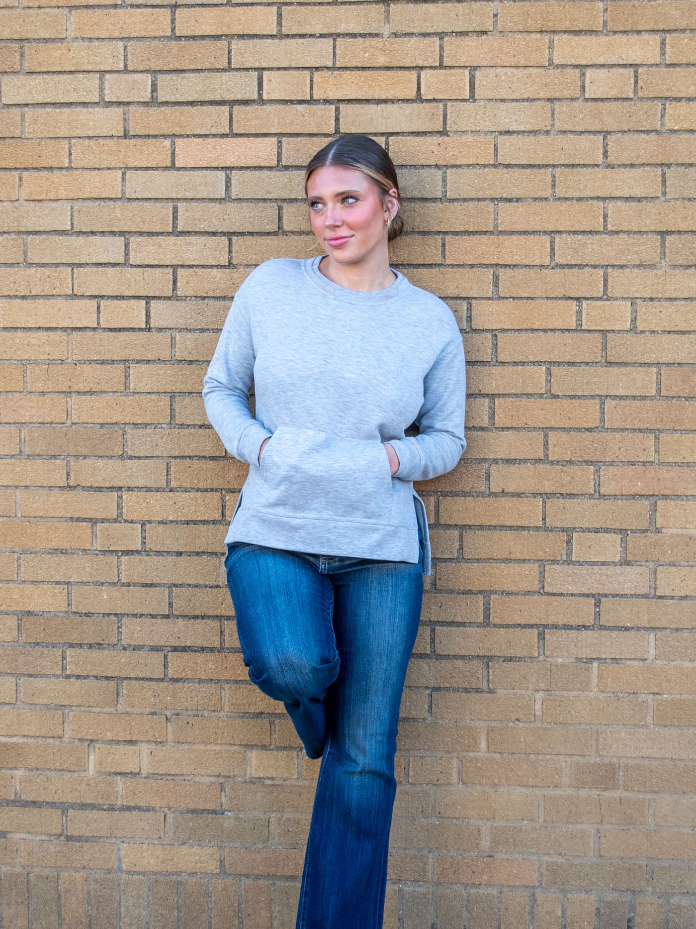 A model wearing a light gray crewneck sweatshirt with a front pocket and side slits. The model has it paired with a dark wash jean.