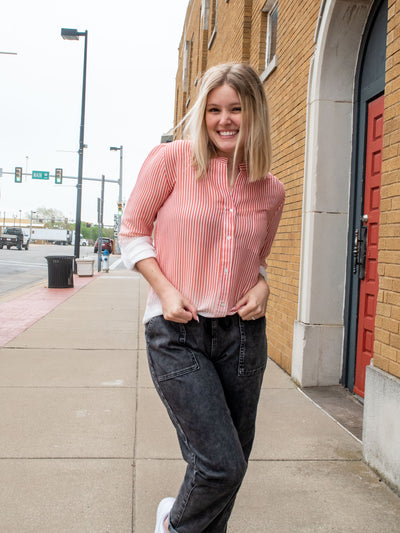 A model wearing a coral to white ombre button down stripe shirt that is long sleeve. The model has it on with washed black denim joggers.