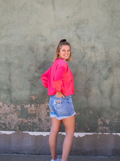 A model wearing a hot pink colored cropped sweatshirt with arm stitching details. She has it on with light washed, frayed jean shorts