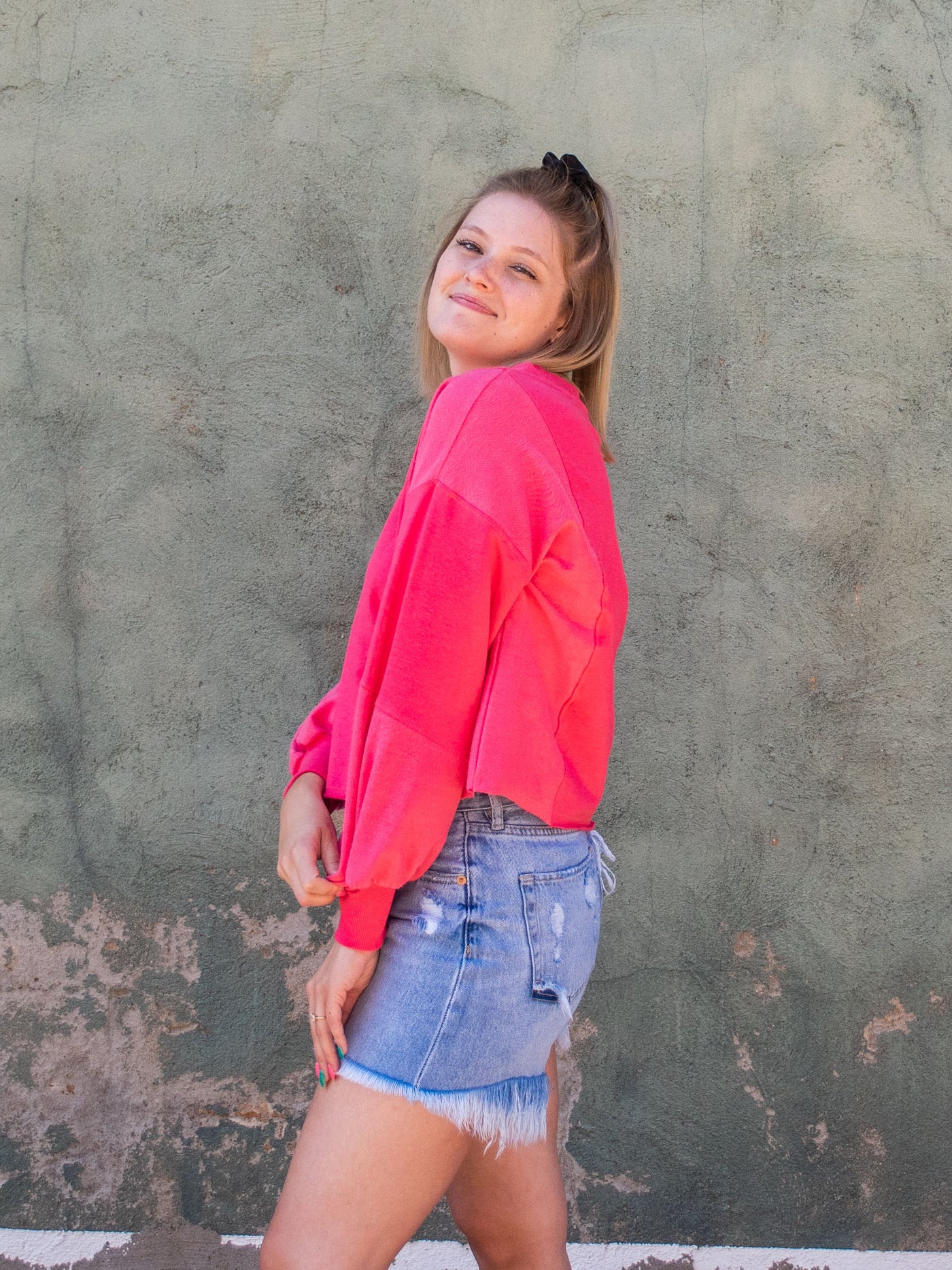 A model wearing a hot pink colored cropped sweatshirt with arm stitching details. She has it on with light washed, frayed jean shorts.