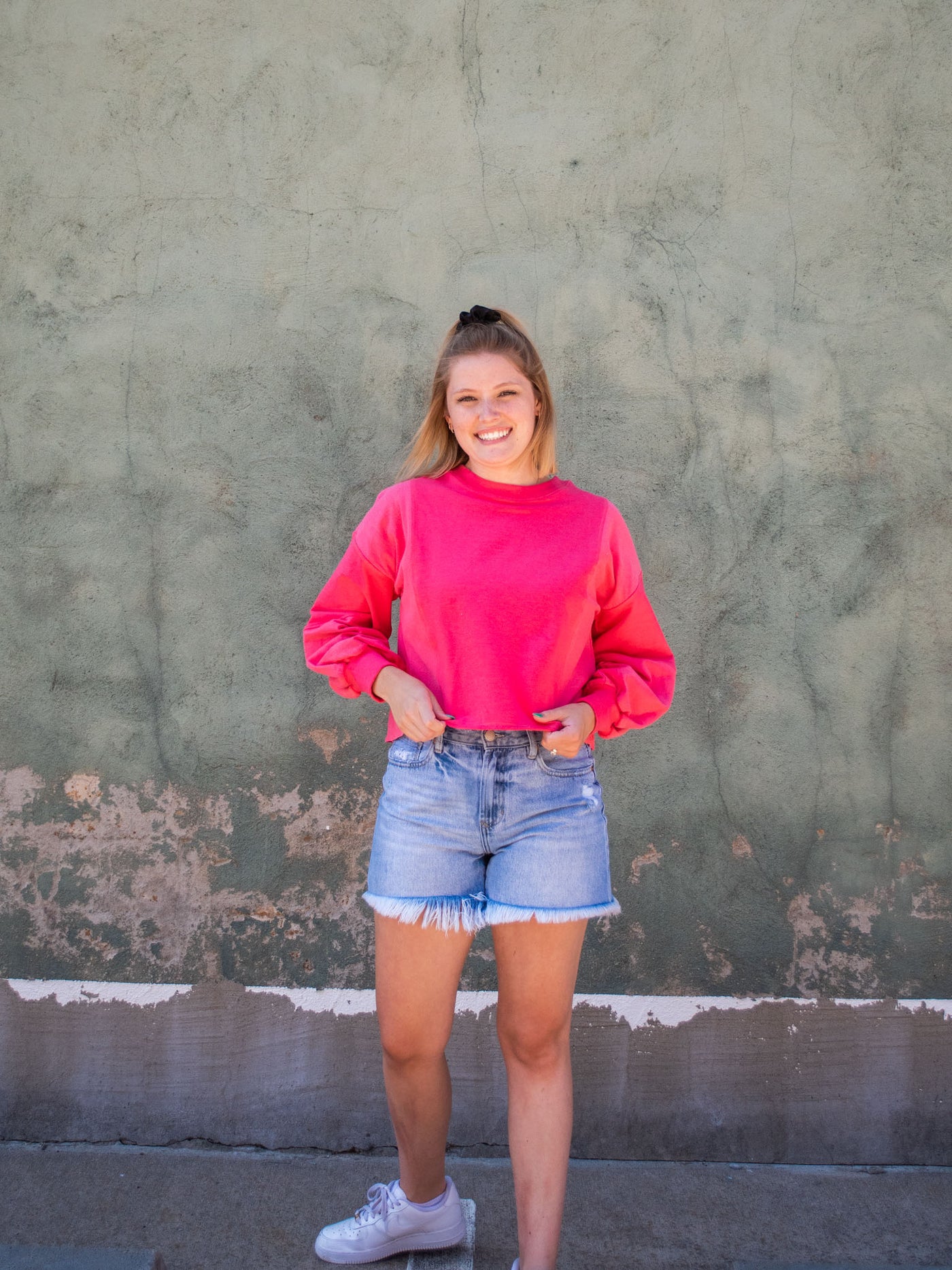A model wearing a hot pink colored cropped sweatshirt with arm stitching details. She has it on with light washed, frayed jean shorts