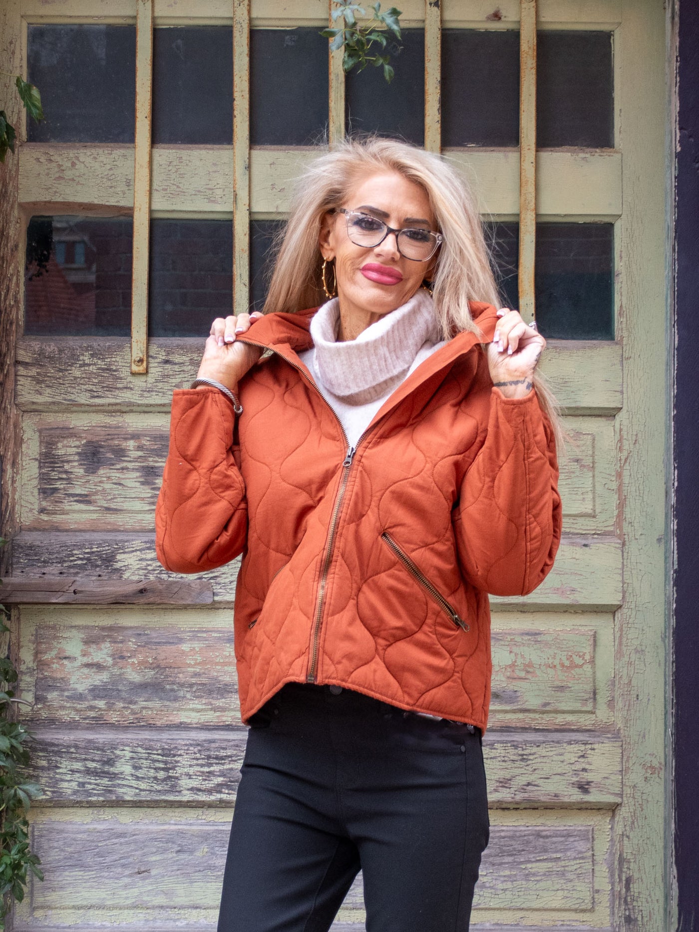 A model wearing a rust collared quilted zip-up jacket. The model has it paired with a cream turtleneck and a black jean.