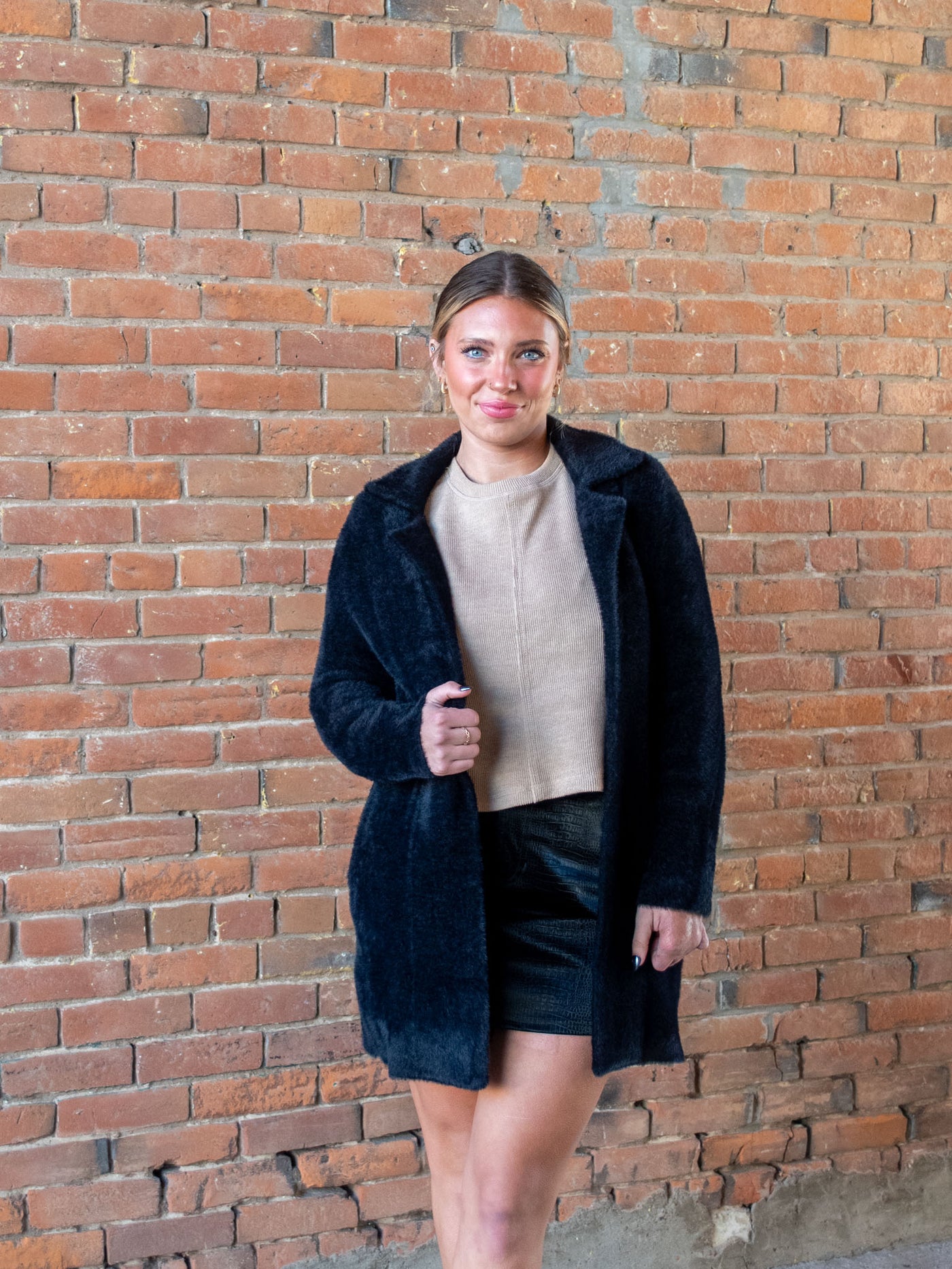 A model wearing a black fuzzy open front coatigan. The model has it paired on top of a tan sweater and a black faux leather mini skirt. 