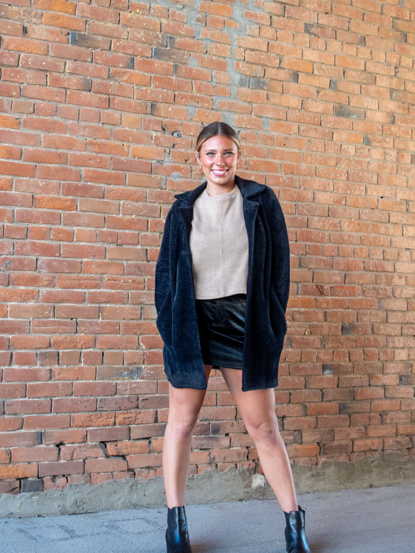 A model wearing a black fuzzy open front coatigan. The model has it paired on top of a tan sweater, a black faux leather mini skirt and black booties. 