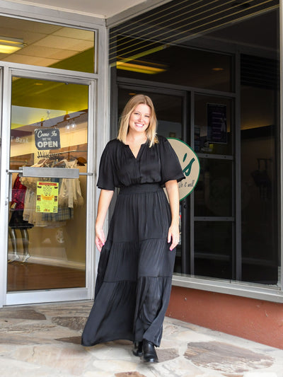 A model wearing a black tiered maxi dress with a long, short sleeve. The model has it paired with a black bootie.