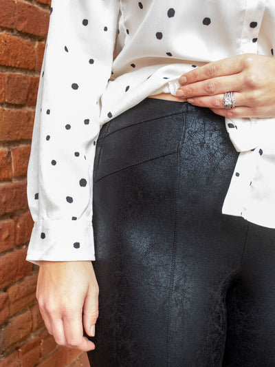 A model wearing a pair of black faux leather crackle leggings. The model has it paired with a white and black polka dot blouse and white heels.