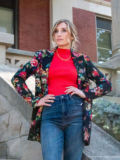 A model wearing a black blazer with a floral print. The model has it paired with a red top underneath and a medium wash jean.