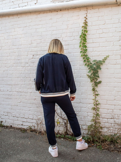 A model wearing a navy zip-up jacket with a striped detail on the neckline, edge, and sleeve cuff. She has it paired with a white top underneath, navy joggers, and white sneakers.