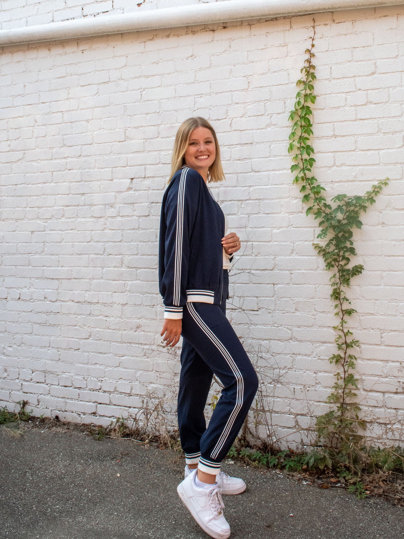 A model wearing a navy zip-up jacket with a striped detail on the neckline, edge, and sleeve cuff. She has it paired with a white top underneath, navy joggers, and white sneakers.
