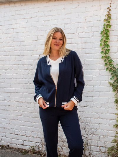 A model wearing a navy zip-up jacket with a striped detail on the neckline, edge, and sleeve cuff. She has it paired with a white top underneath and navy joggers.