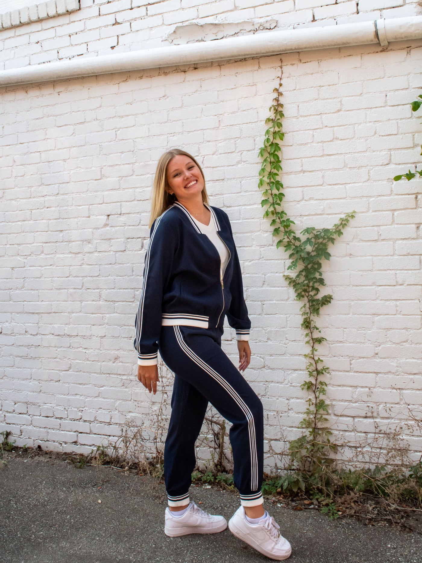 A model wearing a pair of navy joggers with a white and navy stripped cuff on the leg. The model has them paired with a matching navy jacket, a white top, white sneakers.