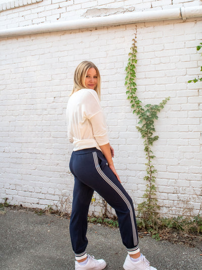 A model wearing a pair of navy joggers with a white and navy stripe down the side and a striped cuff on the leg. The model has them paired with a white top and white sneakers.