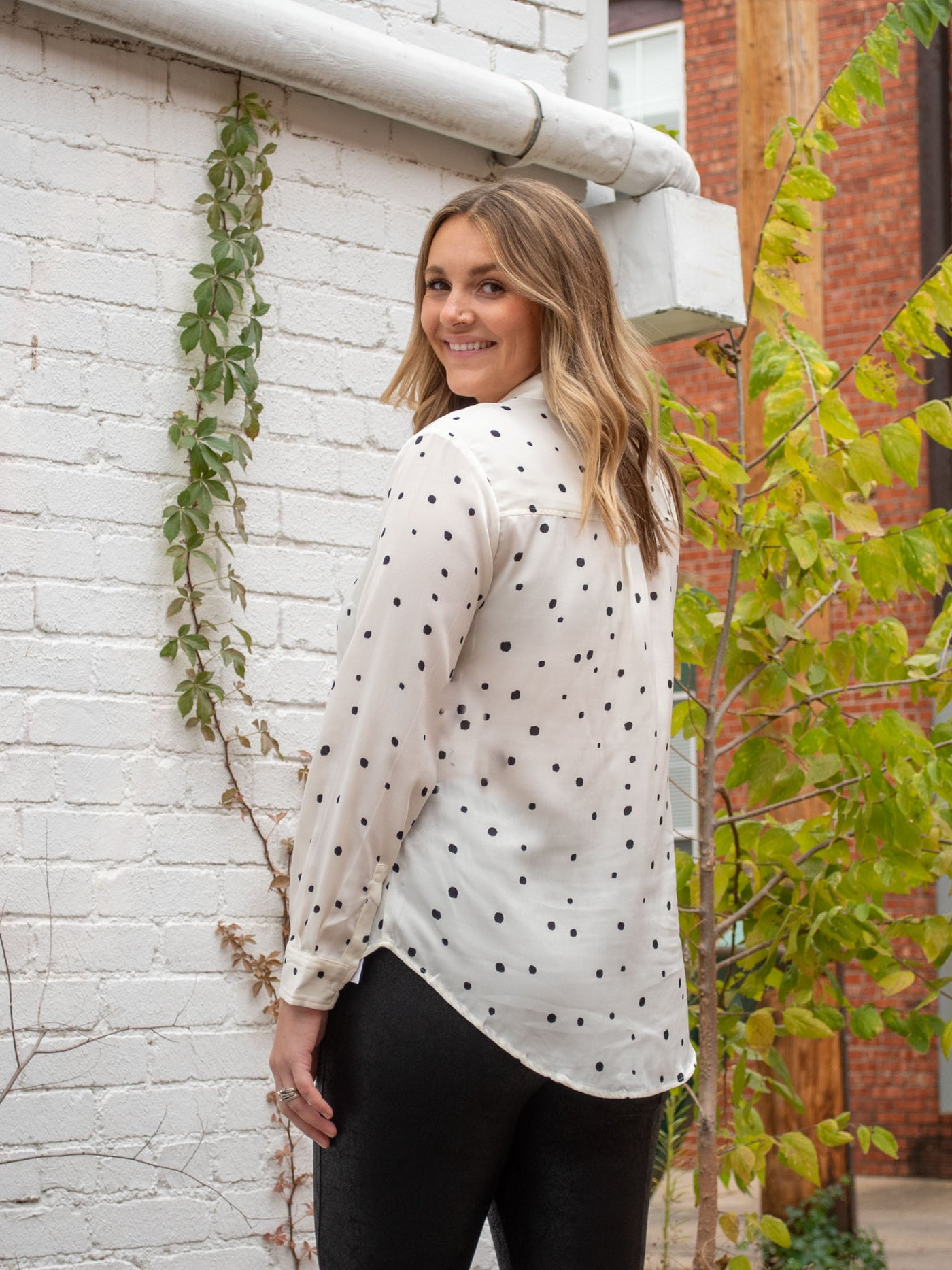 A model wearing a white button down blouse with polka dots. The model has it paired with pleather pants.