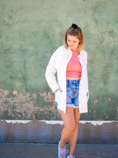 A model wearing a white button down top with pockets. She has it over a pink cropped tank top and light washed jean shorts with white sneakers. 