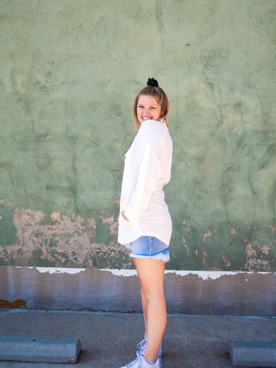 A model wearing a white button down top with pockets. She has it over a pink cropped tank top and light washed jean shorts with white sneakers. 