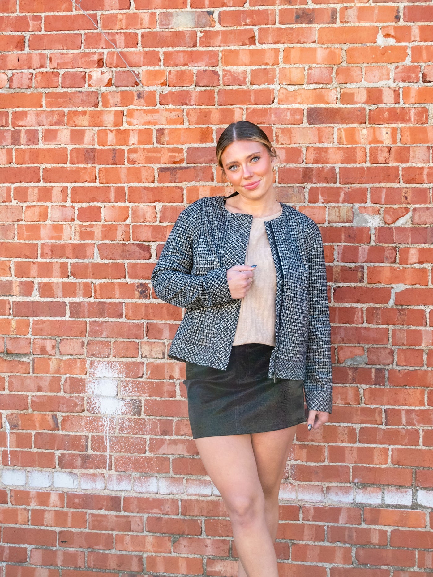 A model wearing a plaid and white small plaid, zip up jacket. The model has it paired with a tan sweater and faux leather mini skirt.
