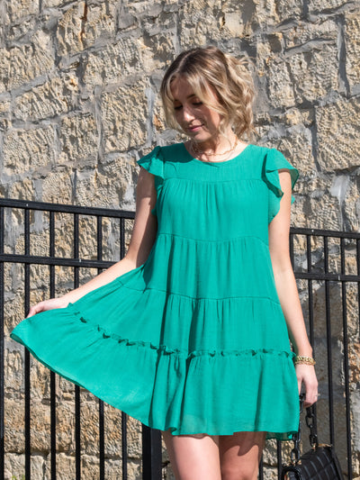 A model wearing a Kelly green ruffled tiered lined mini dress. She has it on with white sneakers.