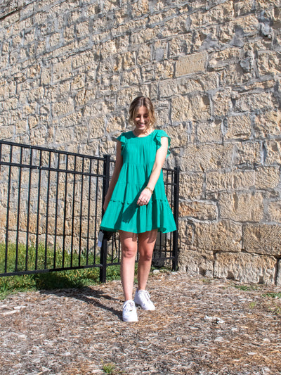 A model wearing a Kelly green ruffled tiered lined mini dress. She has it on with white sneakers.