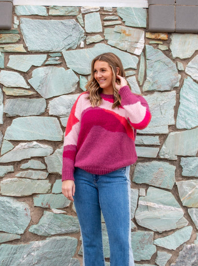 A model wearing a pink, orange, and purple printed crewneck sweater. The model has it paired with two toned jeans.