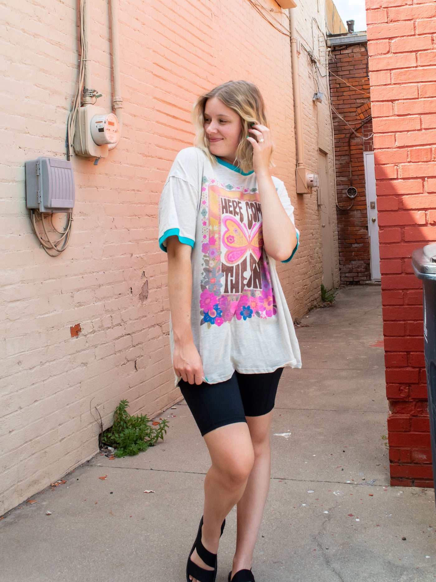 A model wearing an oversized gray graphic tee with contrasting teal blue bands on the sleeves and neckline, and the saying "here comes the sun" surrounded by flowers and butterflies. The model has it paired with black biker shorts and black sandals.