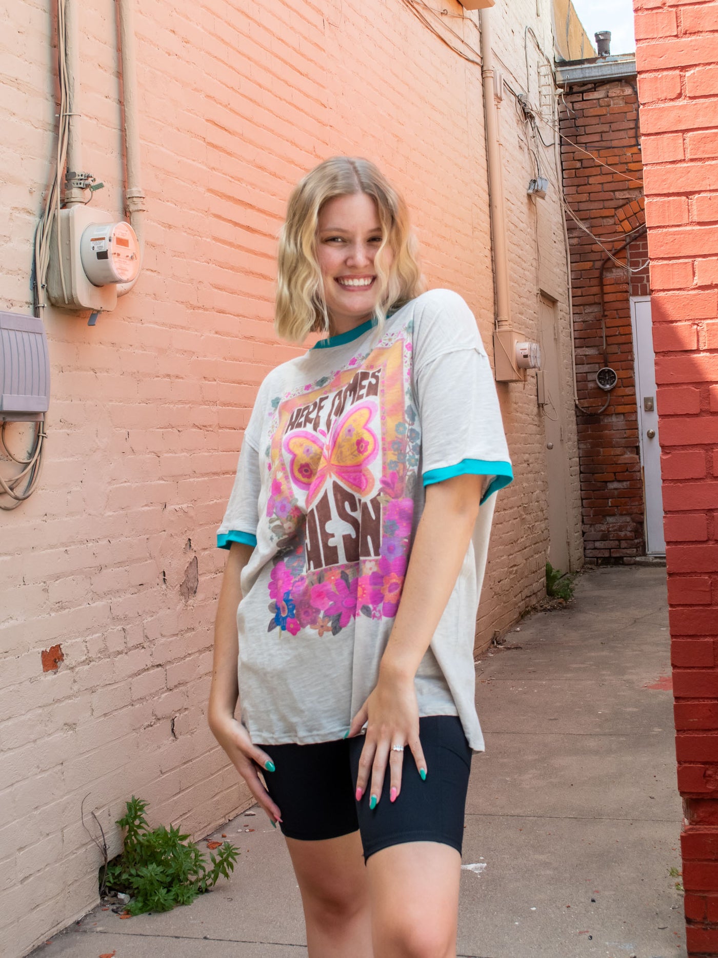 A model wearing an oversized gray graphic tee with contrasting teal blue bands on the sleeves and neckline, and the saying "here comes the sun" surrounded by flowers and butterflies. The model has it paired with black biker shorts.