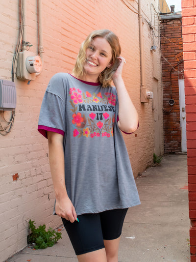 A model wearing an oversized gray graphic tee with contrasting maroon bands on the sleeves and neckline, and the saying "manifest it" surrounded by flowers. The model has it paired with black biker shorts.