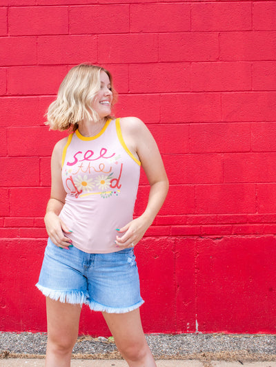 A model wearing a graphic pink tank which says "see the good" with contrasting borders. She has it paired with a pair of frayed denim shorts.