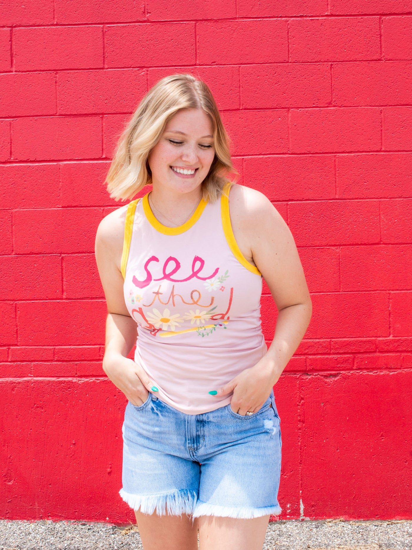 A model wearing a graphic pink tank which says "see the good" with contrasting borders. She has it paired with a pair of frayed denim shorts.