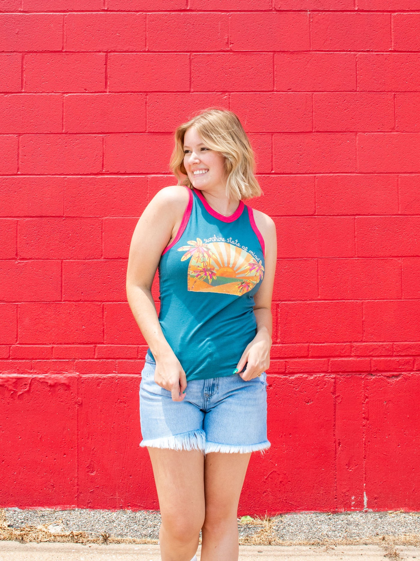 A model wearing a graphic blue fitted tank which says "sunshine state of mind" with contrasting pink borders. She has it paired with a pair of frayed denim shorts.
