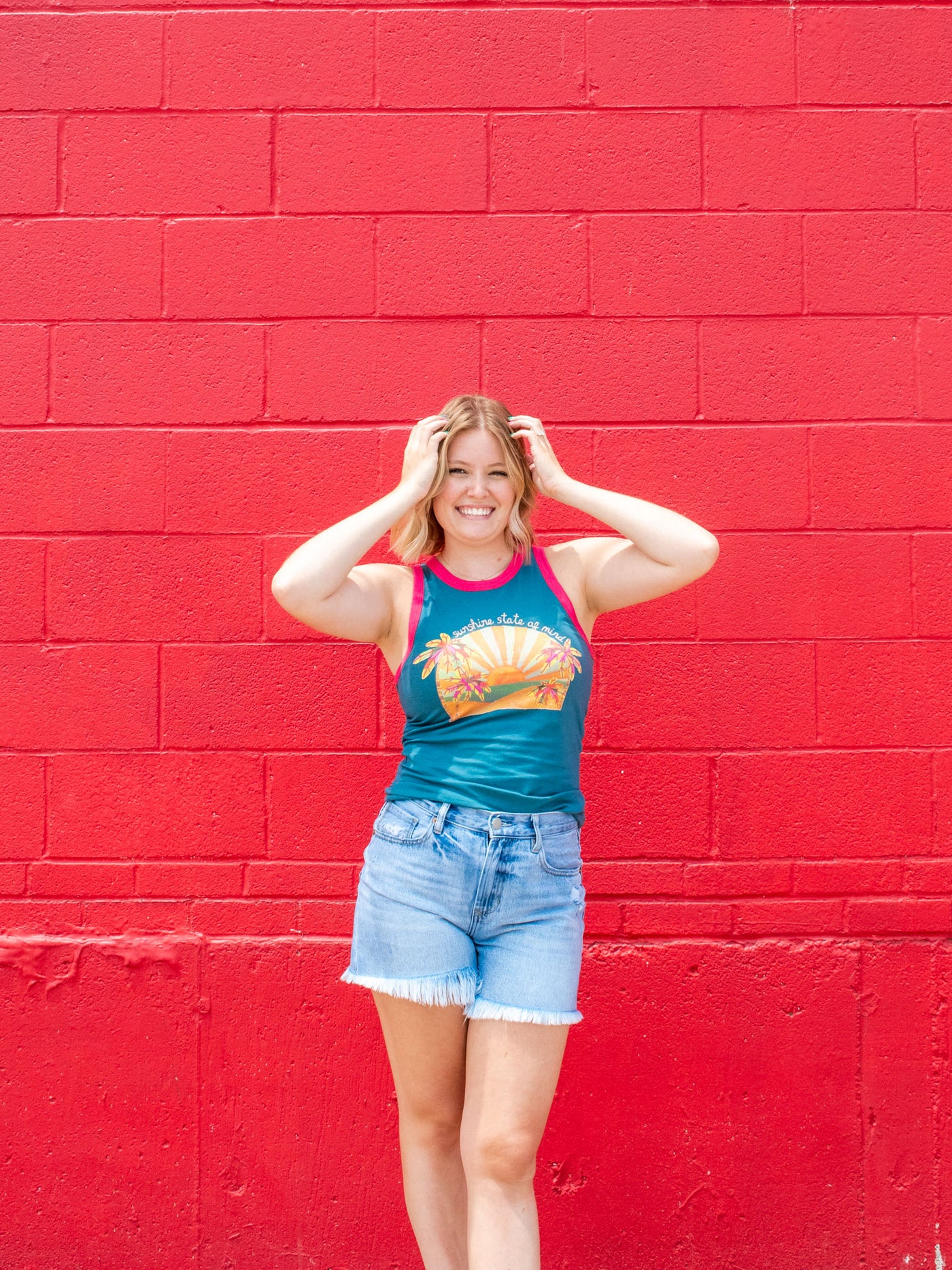 A model wearing a graphic blue fitted tank which says "sunshine state of mind" with contrasting pink borders. She has it paired with a pair of frayed denim shorts.
