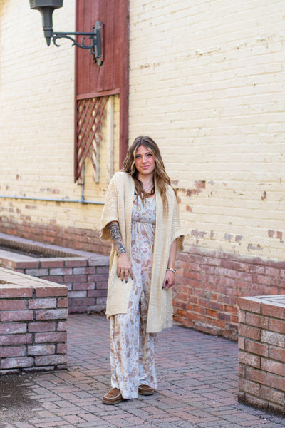 A model wearing a neutral printed jumpsuit. The model has it paired with a chunky tan cardigan and brown loafers.