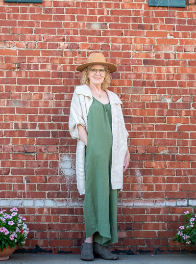 A model wearing an olive green jumpsuit with a v-neckline and pockets. The model has it paired under a chunky knit cardigan, brown hat, and gray booties.
