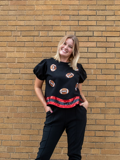 A model wearing a black puff sleeve tee with a red and black wavy hem and sequin footballs. The model has it paired with a black jogger. 