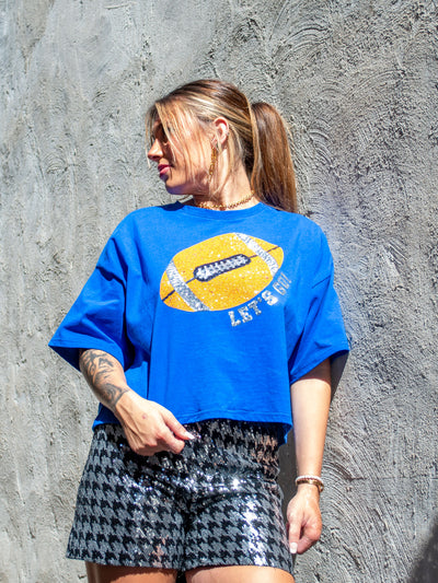 A model wearing a blue boxy tee with a sequin football and the phrase "let's go" on it. She has it paired with black and silver sequin shorts.