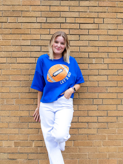 A model wearing a blue boxy tee with a sequin football graphic that says "Let's Go!". The model has it paired with a white wide leg jean.
