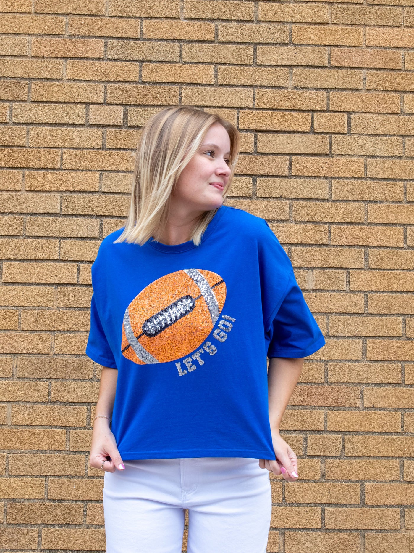 A model wearing a blue boxy tee with a sequin football graphic that says "Let's Go!". The model has it paired with a white wide leg jean.