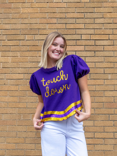 A model wearing a purple top that says touchdown and has purple and gold sequins. The model has it paired with a white jean.